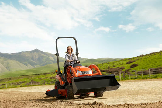 Person operating a Kioti compact tractor in a rural setting with green hills and blue sky.