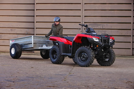 Red and black ATV pulling a trailer with a person standing nearby against a wooden wall.