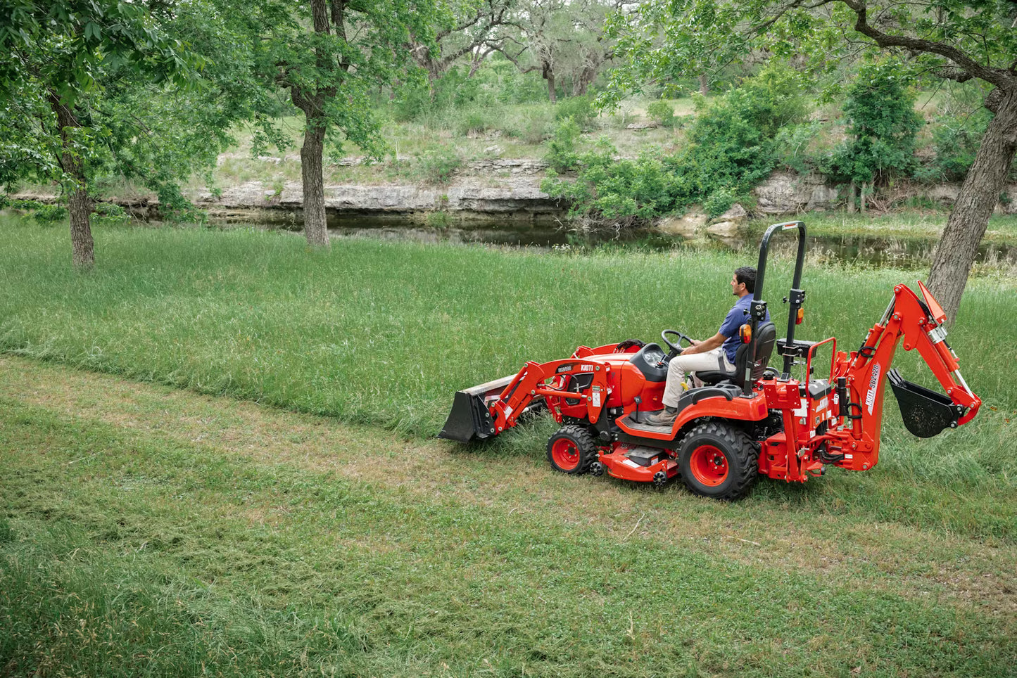 Person operating a Kioti tractor in a grassy outdoor setting with trees and a pond in the background.