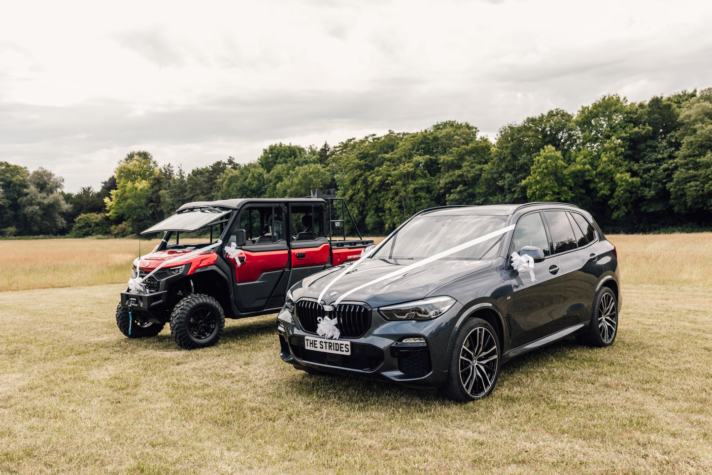 Two wedding cars, one black BMW SUV and one red CFMOTO UFORCE U10 Pro XL, parked on a grassy field with trees in the background.