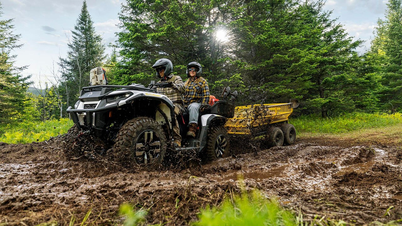 Two people on a Can-Am Outlander Electric ATV with a trailer in a forested area