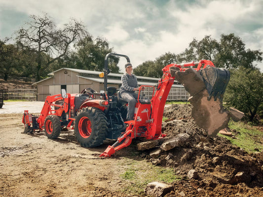 Person operating a Kioti tractor backhoe to break up ground, in a rural setting.
