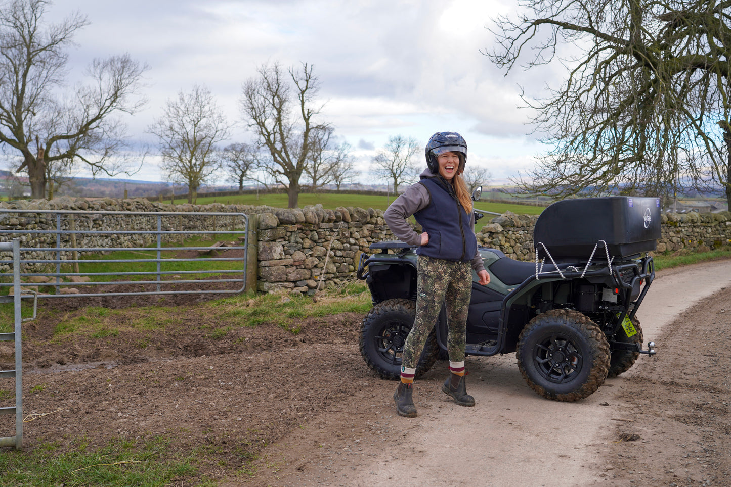Person standing next to a CFMOTO quad bike on a rural road with trees and a stone wall in the background.