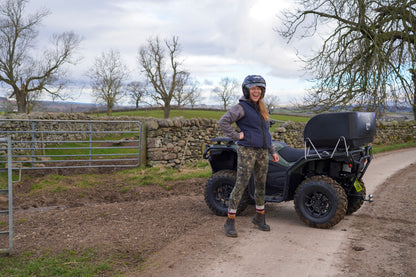 Person standing next to a CFMOTO quad bike on a rural road with trees and a stone wall in the background.