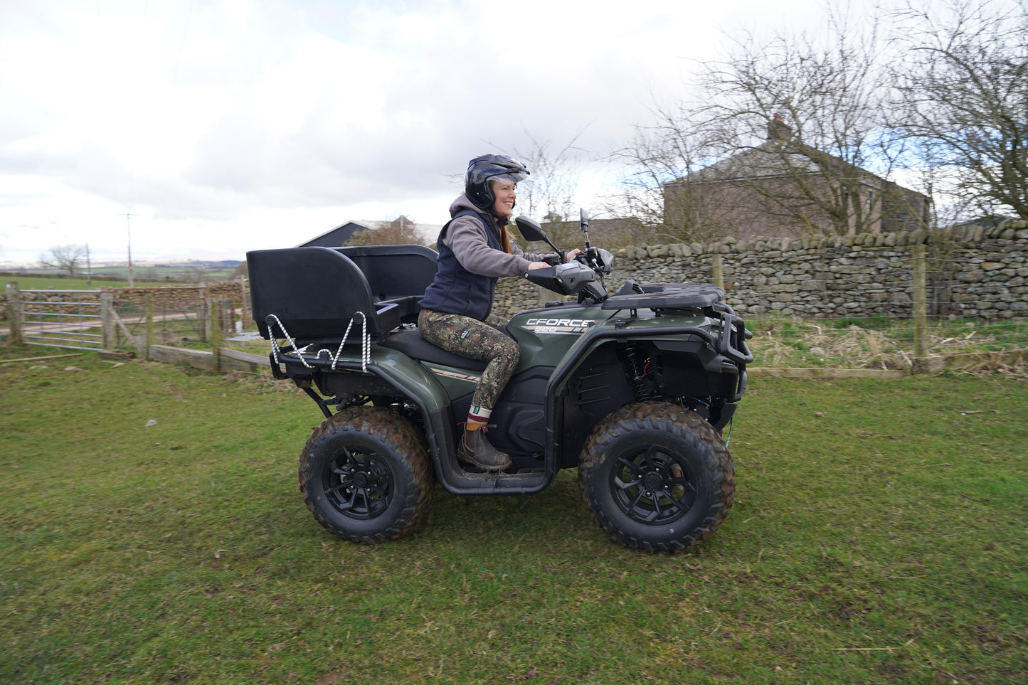Person riding a CFMOTO ATV in a rural setting with a stone wall and wooden fence in the background.