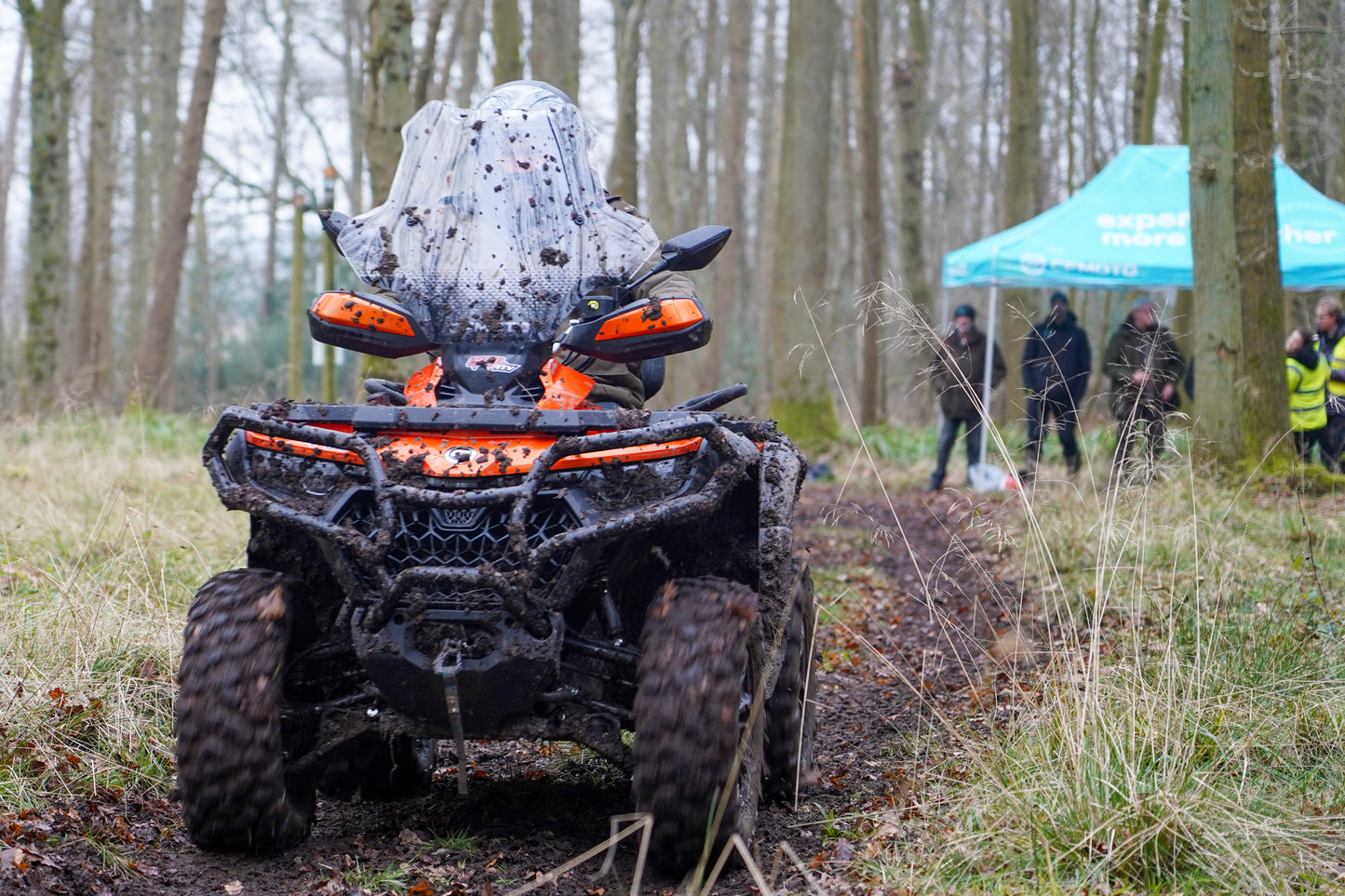 CFMOTO ATV in a forest setting, with people and a tent in the background.