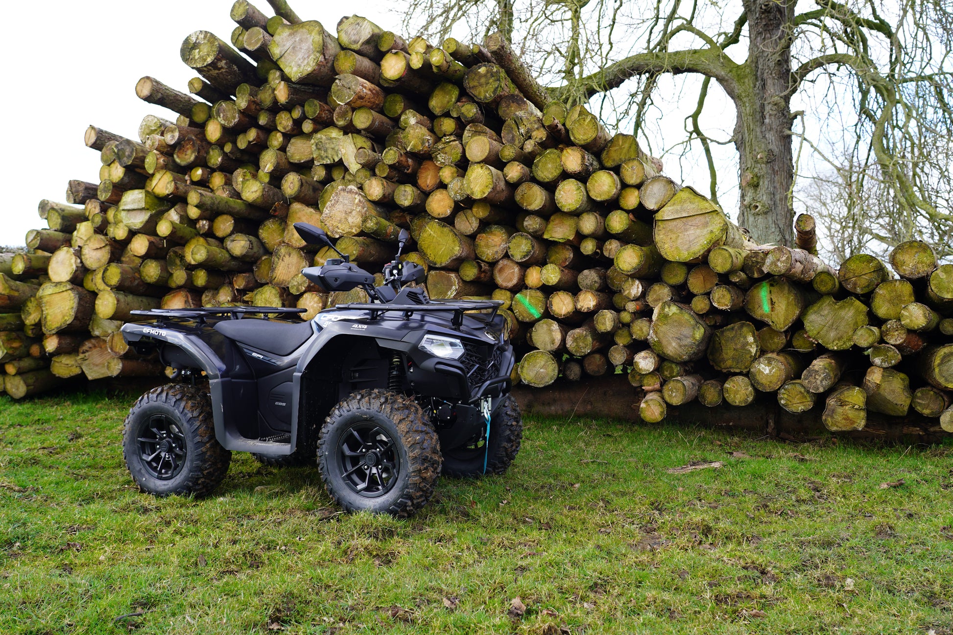 Black CFMOTO ATV parked in front of a large pile of logs in a forest setting