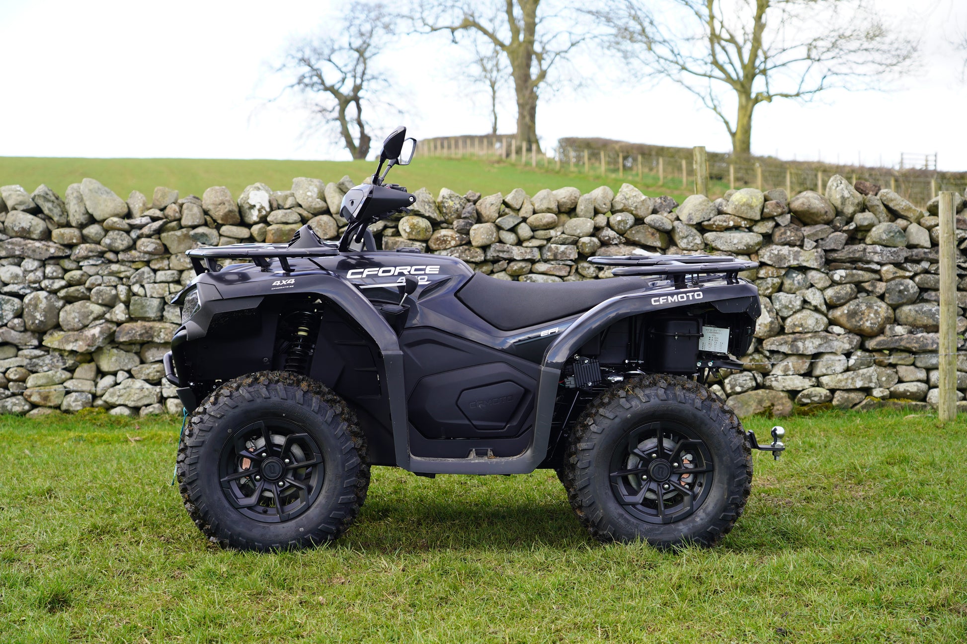 Black CFMOTO quad bike parked in a grassy field with a stone wall and trees in the background