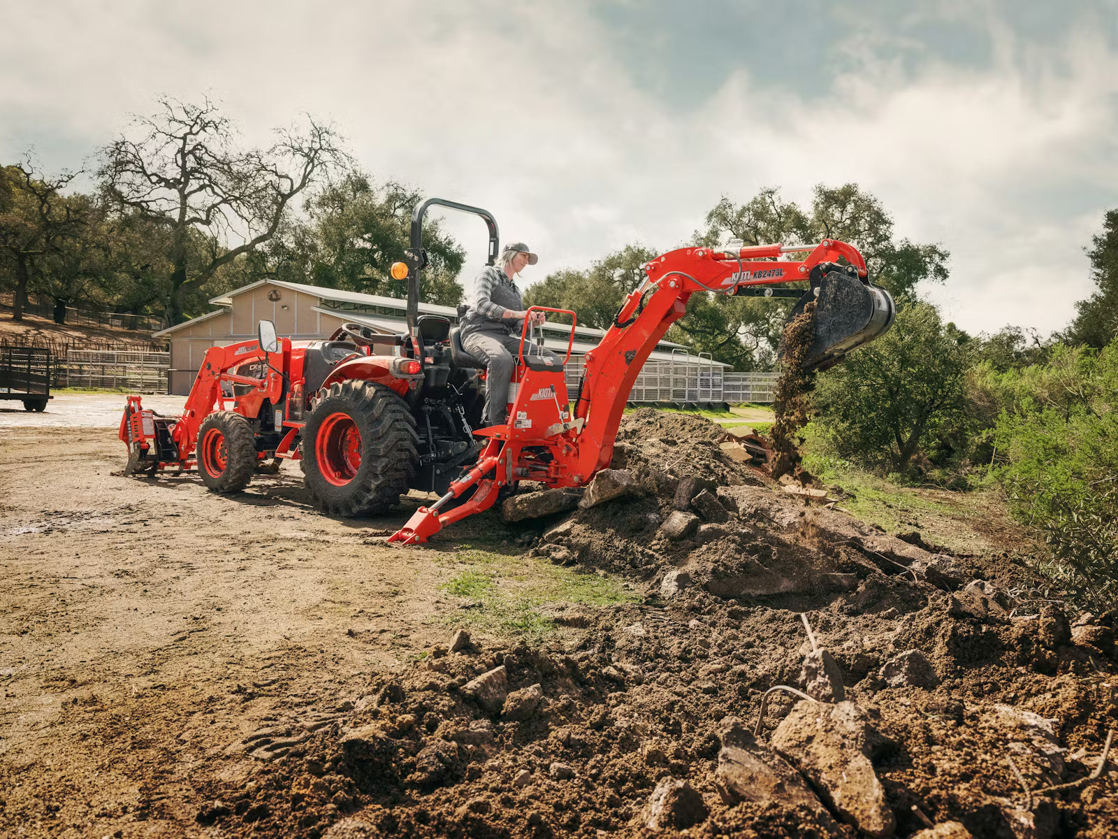 Kioti tractor with an attached backhoe digging in a field with trees and a building in the background.