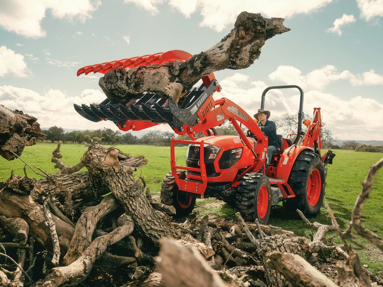 Red Kioti tractor lifting a large log in a field with a person operating it.
