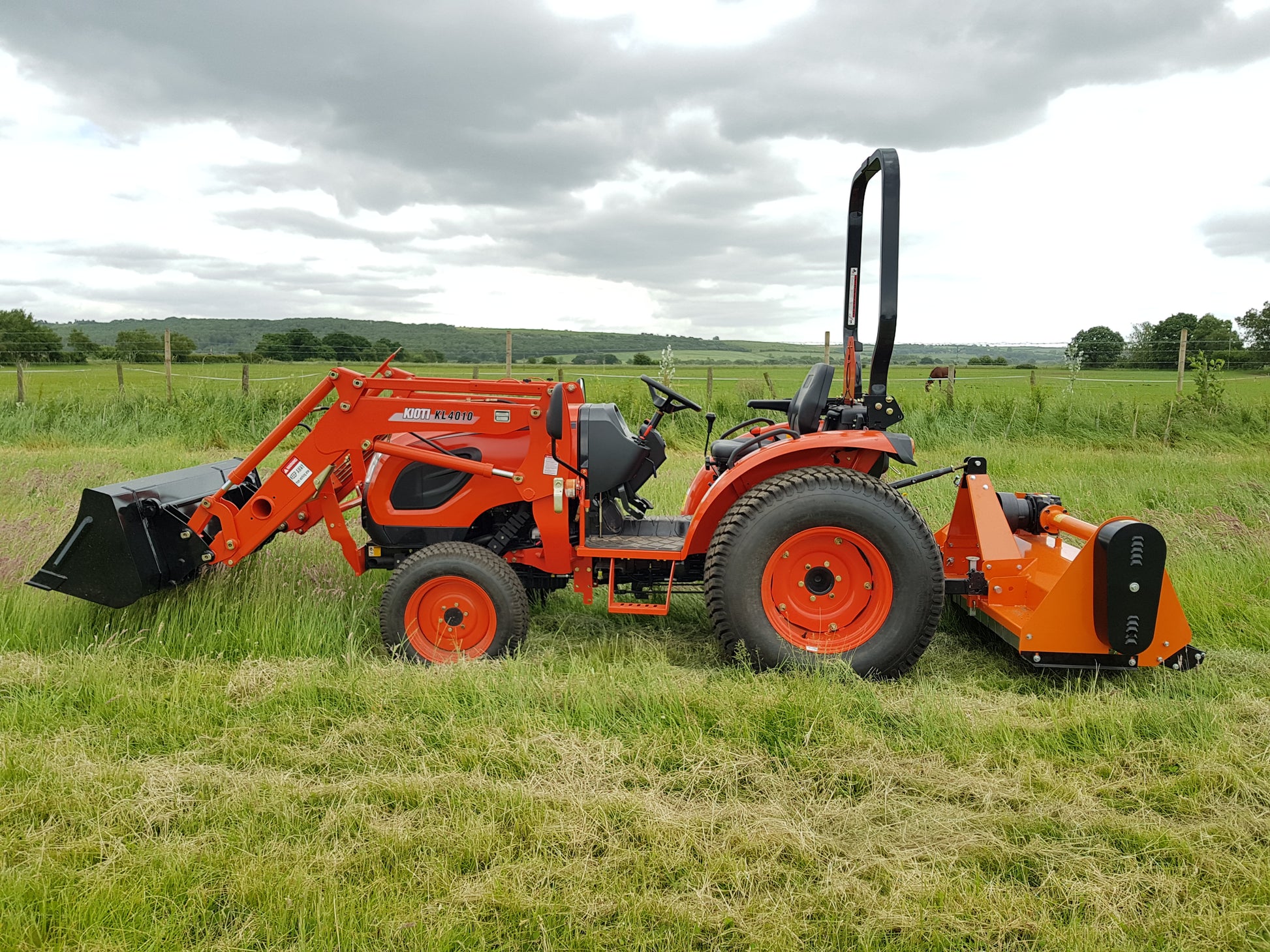 Kioti tractor with flail attachment in a grassy field under a cloudy sky