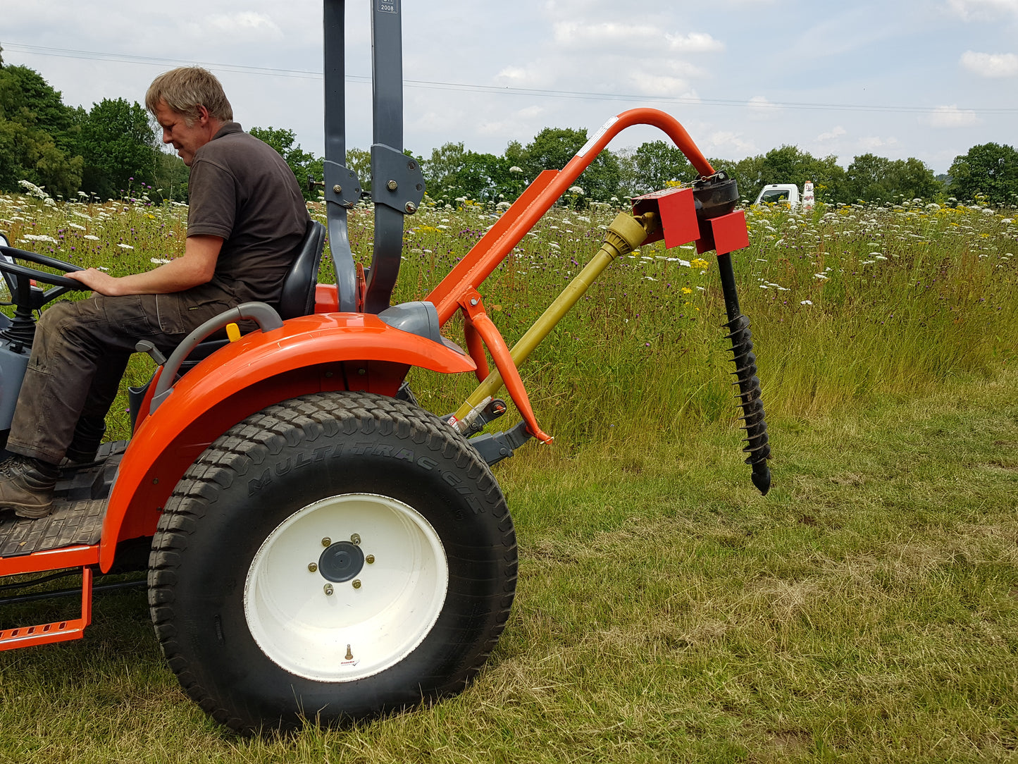 Person operating a small orange tractor with a FarmMaster tractor post hole borer attachment in a field.