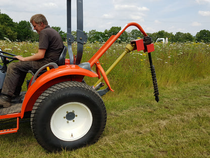 Person operating a small orange tractor with a FarmMaster tractor post hole borer attachment in a field.