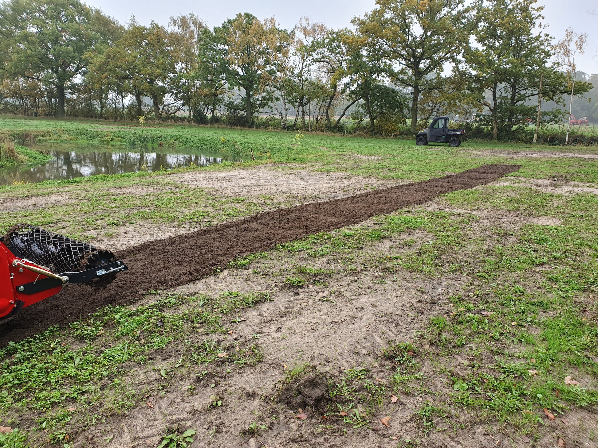 Red tractor with a FarmMaster tractor stone burier in a field with trees and water body in the background