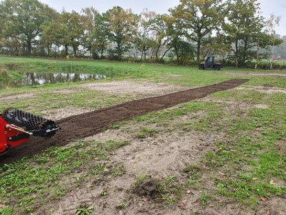 Red tractor with a FarmMaster tractor stone burier in a field with trees and water body in the background