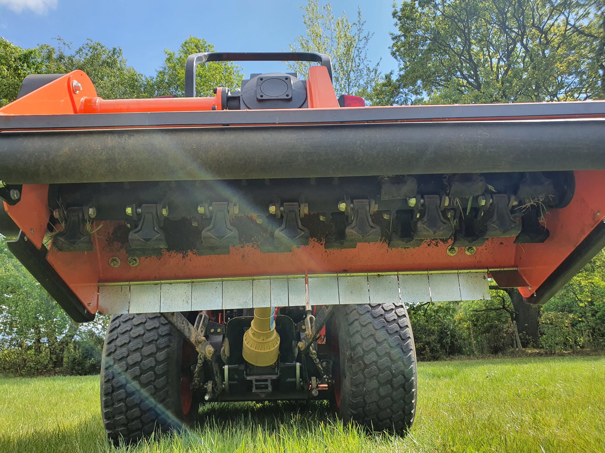 Close-up of a FarmMaster offset flail mower attachment on a grassy field with trees in the background