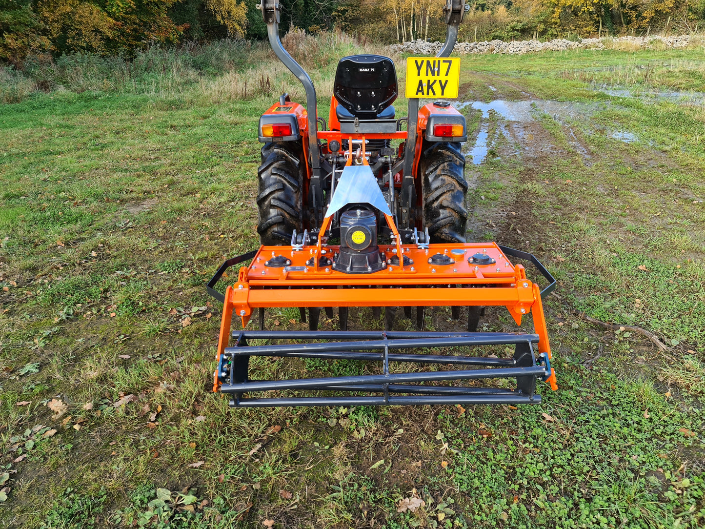 Small orange tractor with a FarmMaster power harrow on a grassy field.