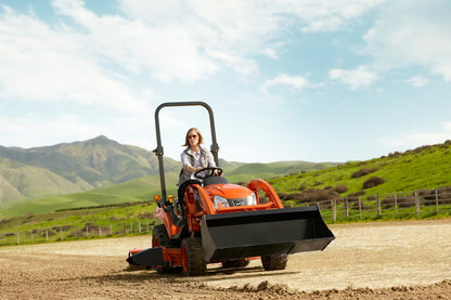 Person operating a Kioti compact tractor in a rural setting with green hills and blue sky.