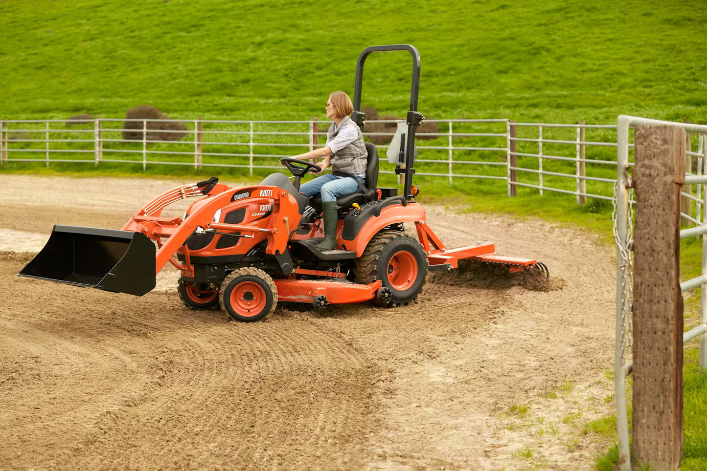 Person operating a Kioti compact tractor in a horse arena with green grass and a fence.