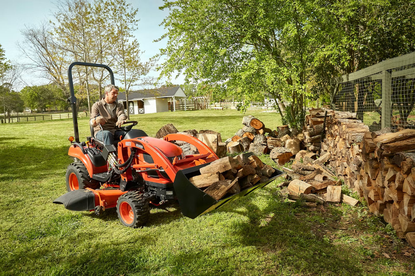 Man operating a Kioti tractor next to stacked firewood in a backyard setting.