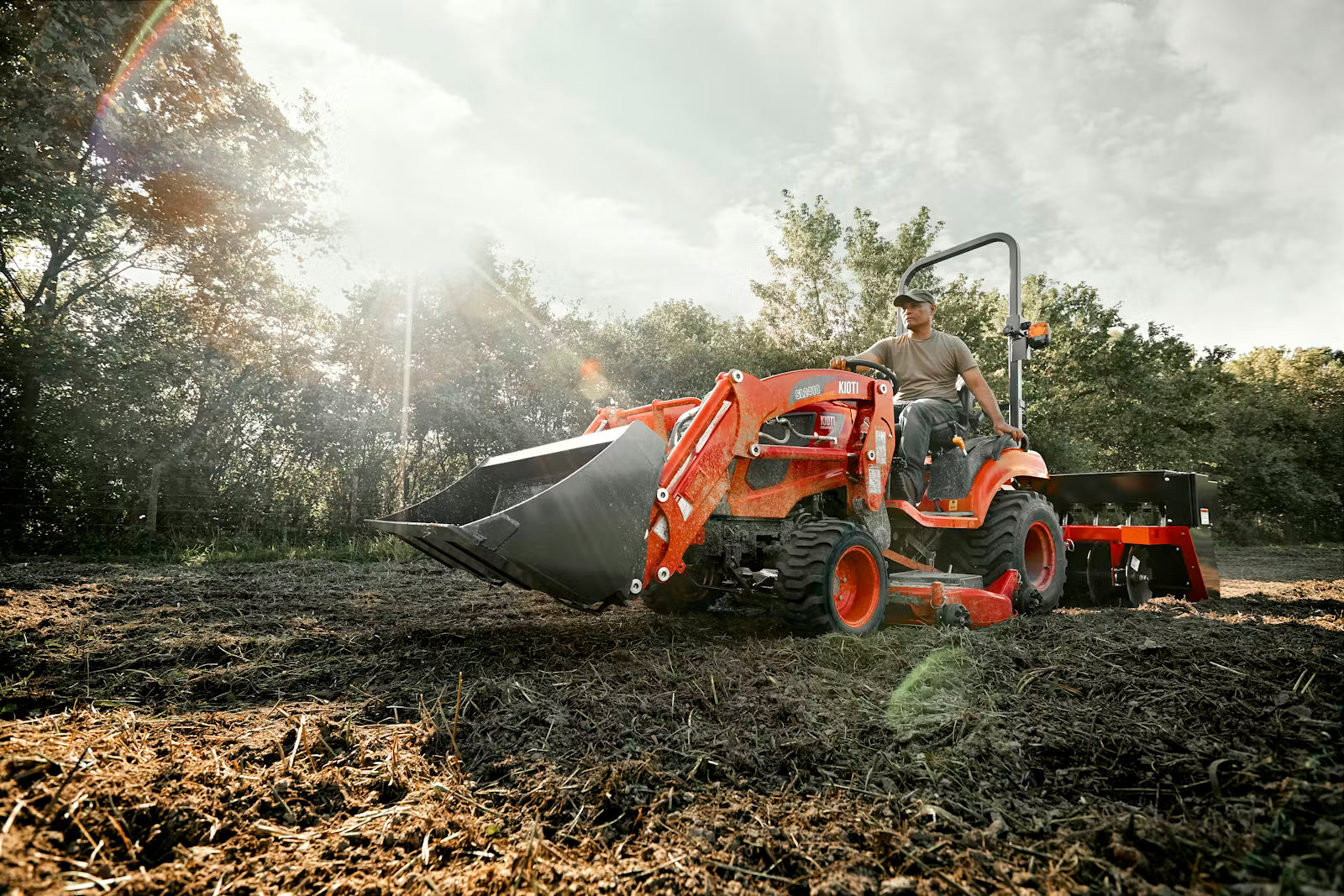 Person operating a Kioti tractor with attachment in a field with trees in the background