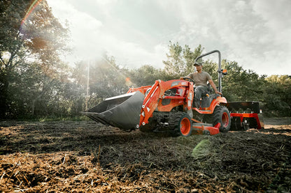 Person operating a Kioti tractor with attachment in a field with trees in the background