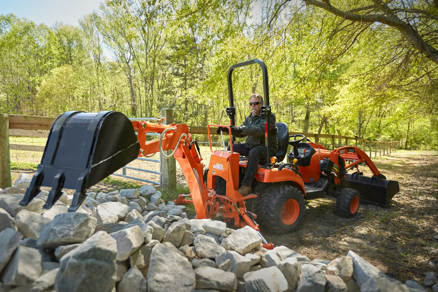Person operating a tractor backhoe on a Kioti tractor in a wooded area with rocks and trees.