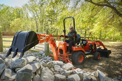 Person operating a tractor backhoe on a Kioti tractor in a wooded area with rocks and trees.