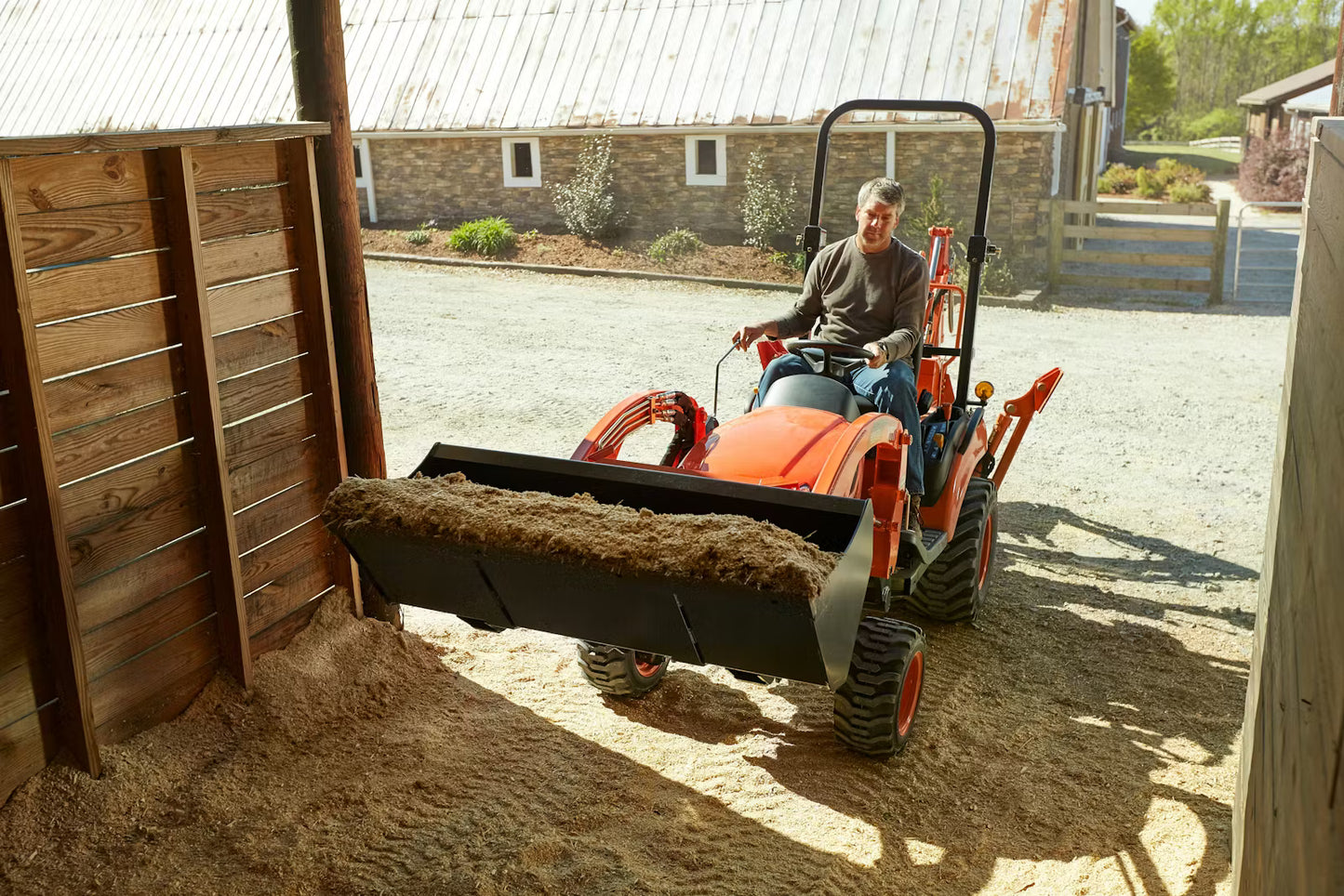 Person operating a small Kioti tractor in an outdoor setting with a wooden structure and barn in the background.