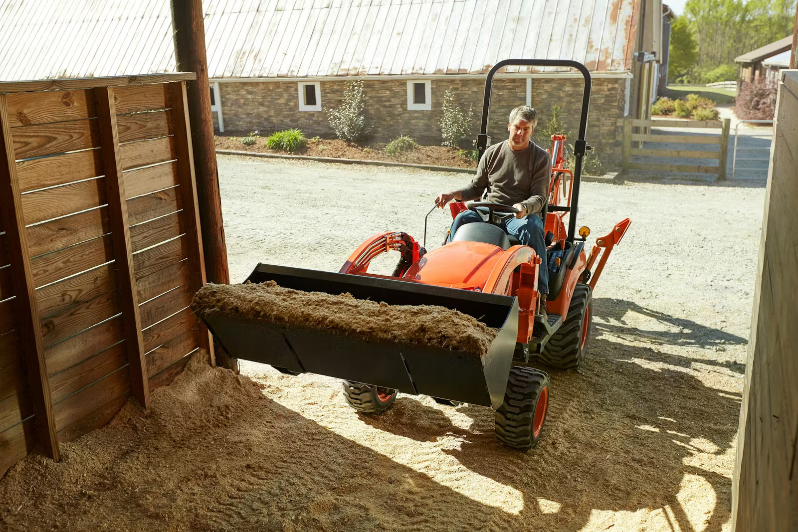 Person operating a small Kioti tractor in an outdoor setting with a wooden structure and barn in the background.