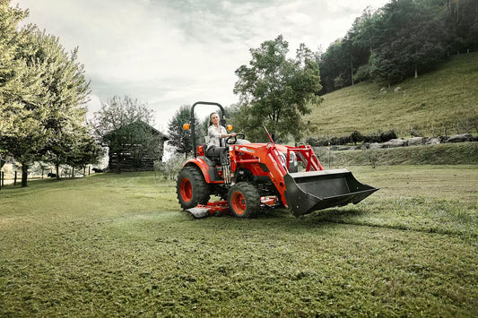 Person operating a Kioti tractor in a grassy field with trees and a house in the background
