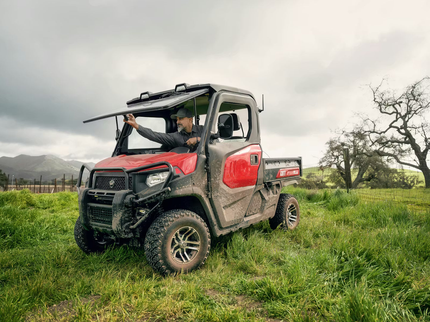 Person driving a Kioti K9 off-road vehicle in a grassy field with mountains in the background.