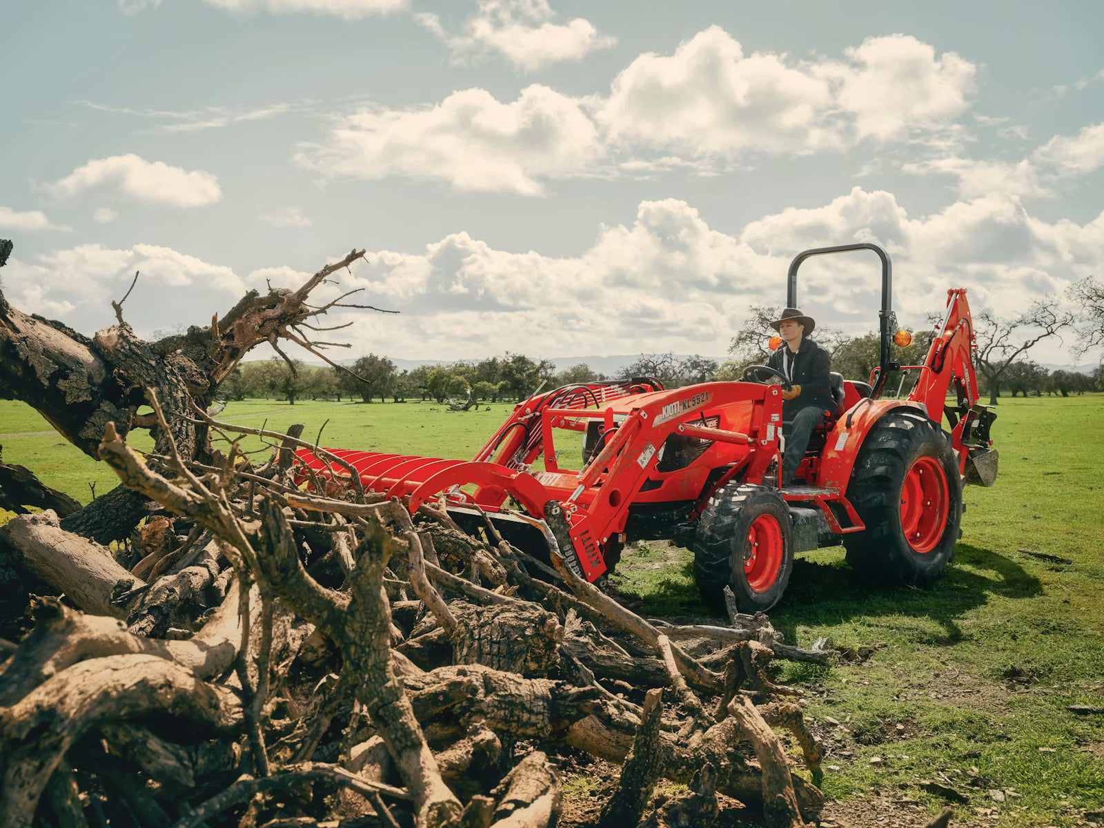 Red Kioti tractor with a backhoe attachment in a field with trees and a person sitting on it.