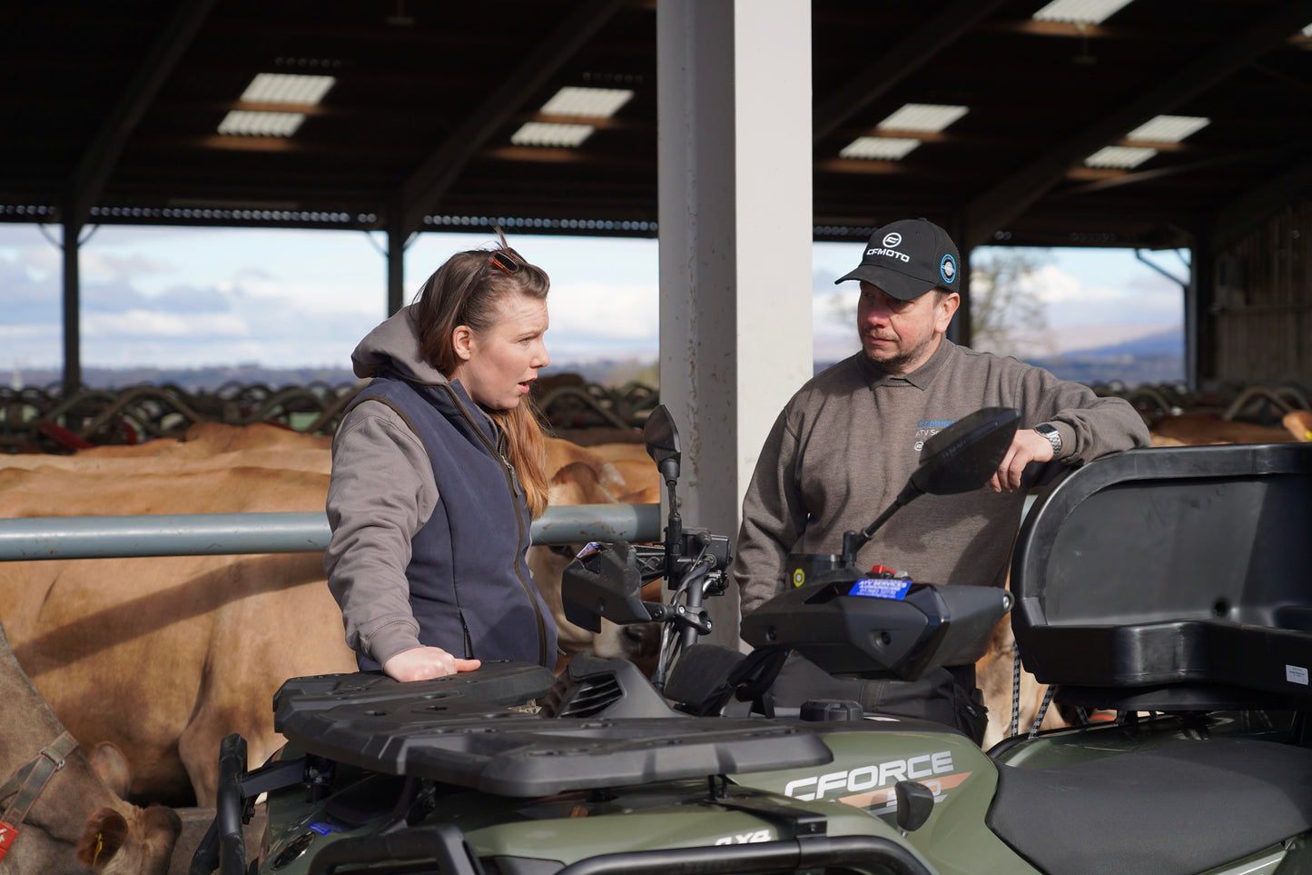Two people with a CFMOTO four-wheeler in a barn setting with cows in the background