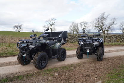 Two CFMOTO quad bikes on a dirt road with a rural landscape in the background.