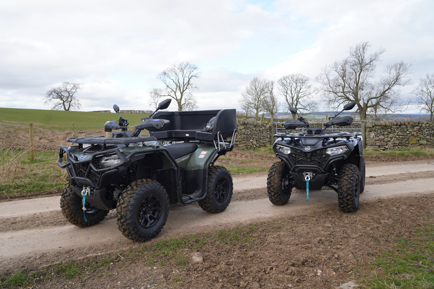 Two CFMOTO all-terrain vehicles on a dirt road with a rural landscape in the background.