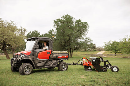 Kioti K9 Utility vehicle towing a stump grinder in a grassy field with trees in the background
