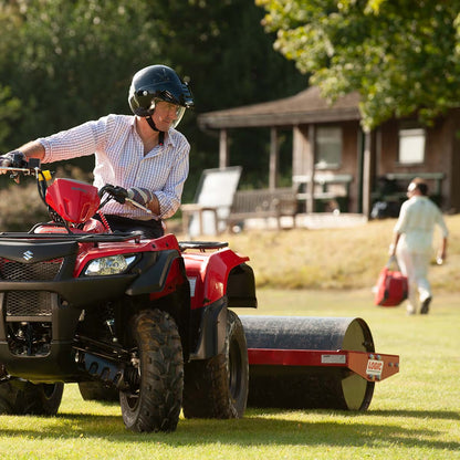 Person operating a red ATV with a logic land roller attachment on a grassy area.