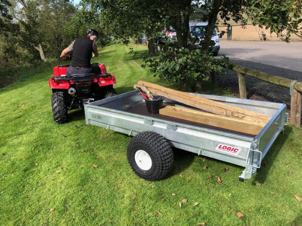 Person on a red ATV with a trailer loaded with wood and tools in a grassy area.