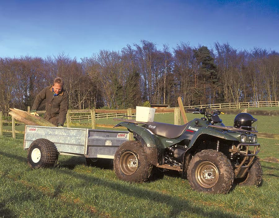 Man loading a trailer onto an ATV in a grassy field with trees in the background
