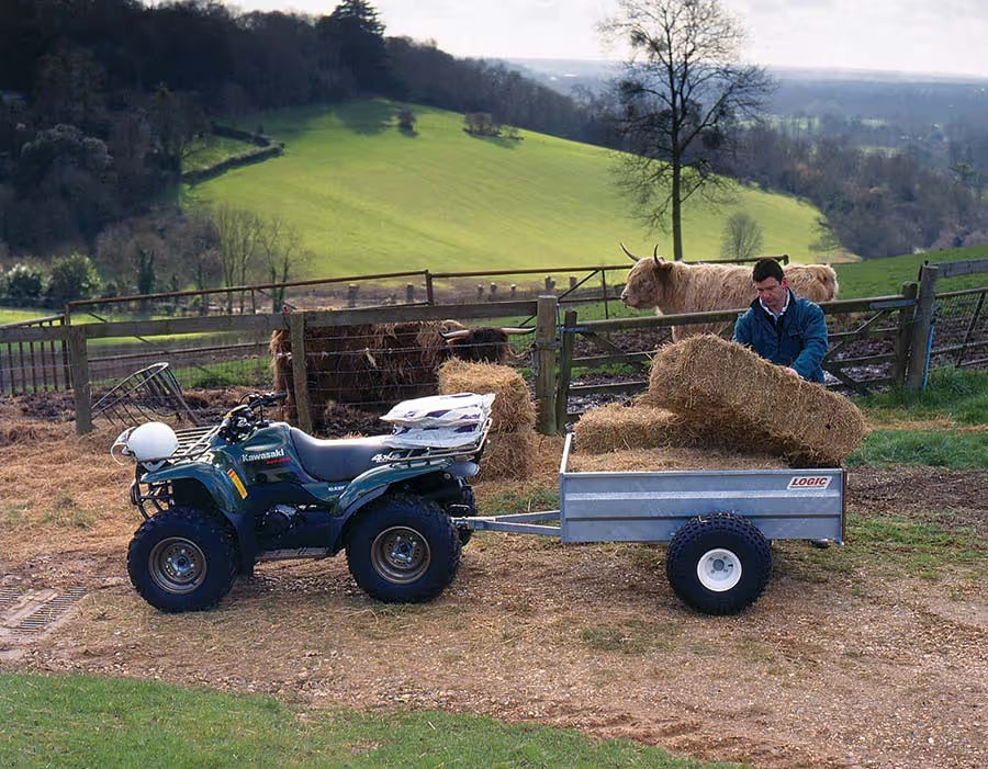 Person with an ATV pulling a trailer loaded with hay near a fenced area with livestock.