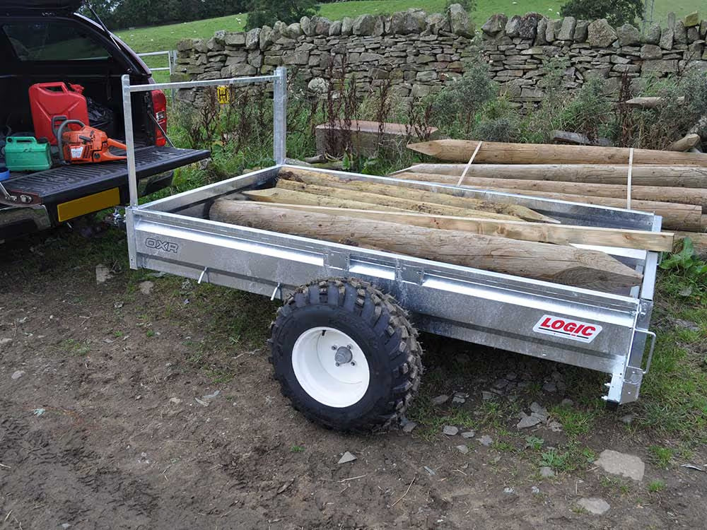 Logic ATV Trailer with wooden logs on a dirt road next to a stone wall.