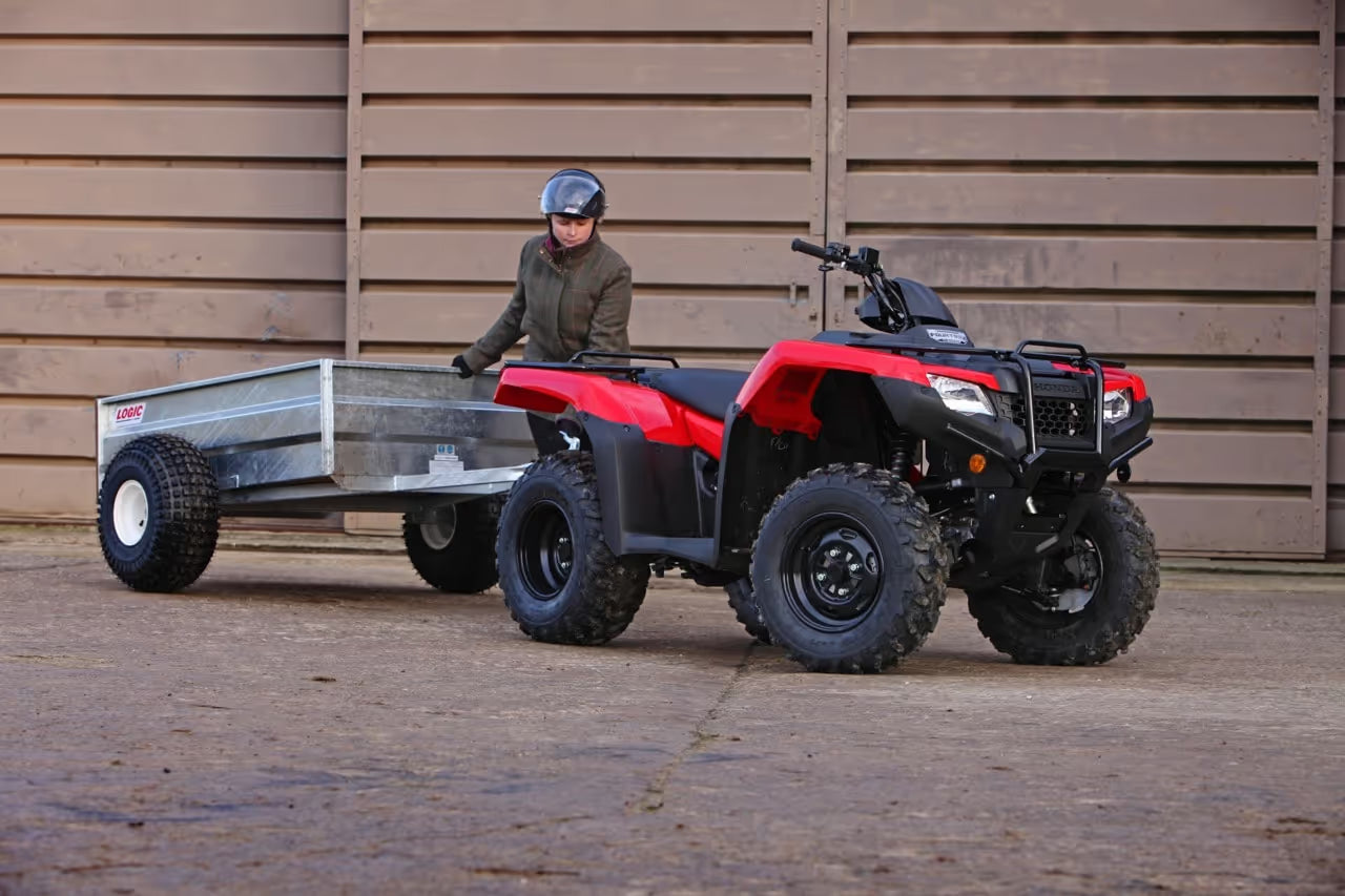 Red and black ATV pulling a trailer with a person standing nearby against a wooden wall.