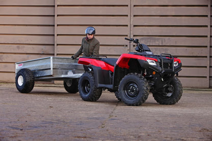 Red and black ATV pulling a trailer with a person standing nearby against a wooden wall.
