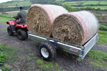 Person on a red ATV pulling a trailer with hay bales in a rural setting