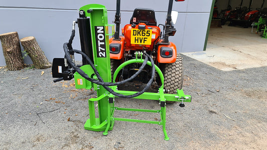 Green FarmMaster Goliath tractor log splitter attached to an orange Kioti tractor in a workshop setting.