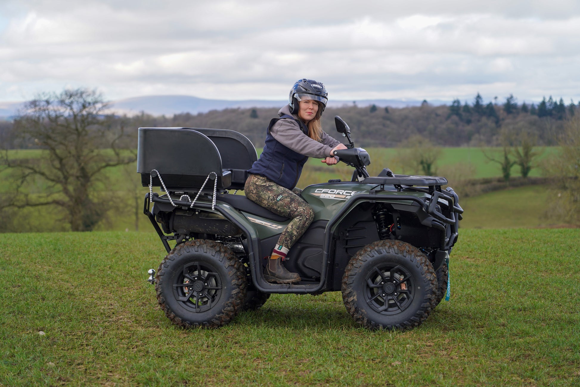 Person riding a green CFMOTO ATV in a grassy field with trees and mountains in the background