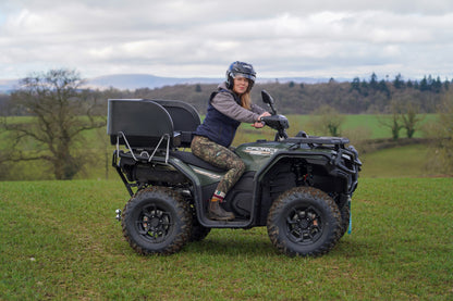 Person riding a green CFMOTO ATV in a grassy field with trees and mountains in the background