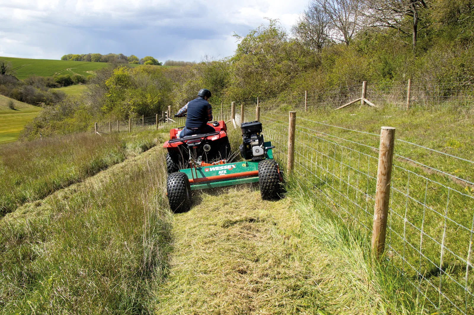 Person operating a rWessex ATV flail mower on a grassy hillside with a fence and trees in the background.