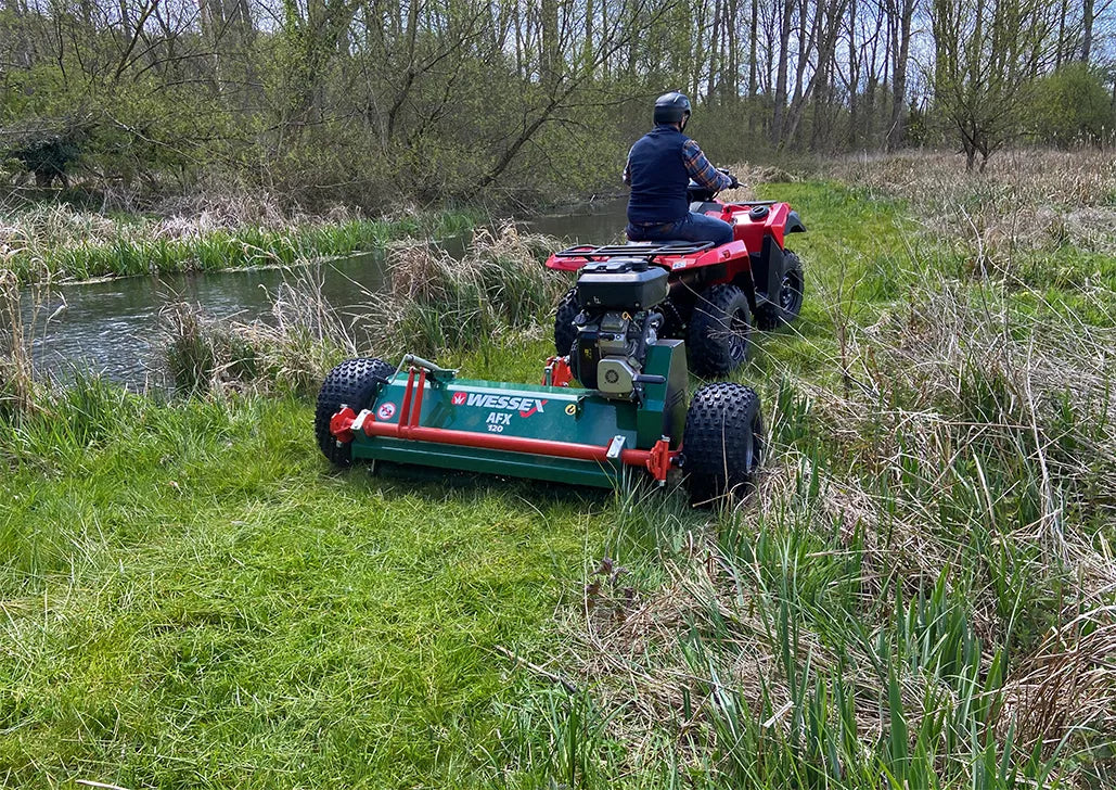 Person operating a large lWessex ATV flail mower on a grassy area with a river in the background
